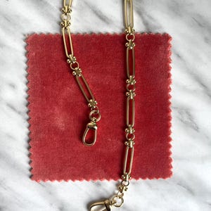 May include: A gold-colored chain with rectangular links and decorative accents. The chain has two clasps and is displayed on a red, textured surface. The chain's design features small, ornate details between the links.