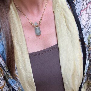 Pode incluir: Um colar de corrente dourada com um pingente em forma de frasco de cristal facetado verde-claro com tampa met&aacute;lica. O colar &eacute; usado sobre uma blusa castanha com um len&ccedil;o amarelo-claro e um casaco com uma estampa semelhante a um mapa.