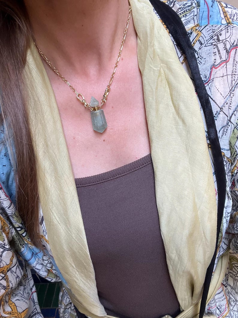 Pode incluir: Um colar de corrente dourada com um pingente em forma de frasco de cristal facetado verde-claro com tampa met&aacute;lica. O colar &eacute; usado sobre uma blusa castanha com um len&ccedil;o amarelo-claro e um casaco com uma estampa semelhante a um mapa.