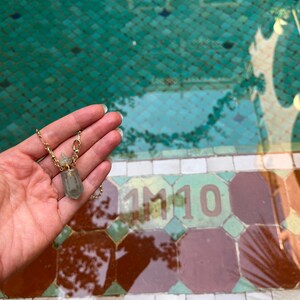 Pode incluir: Um colar com pendente composto por um pequeno frasco de cristal hexagonal transl&uacute;cido em verde p&aacute;lido, com uma tampa de metal dourado e uma corrente a condizer. O frasco foi concebido para conter uma pequena quantidade de l&iacute;quido ou &oacute;leo.