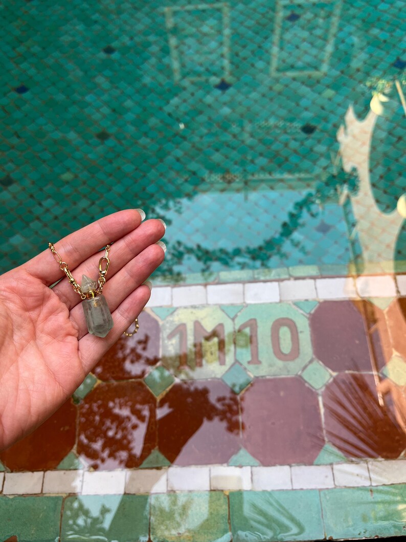 Pode incluir: Um colar com pendente composto por um pequeno frasco de cristal hexagonal transl&uacute;cido em verde p&aacute;lido, com uma tampa de metal dourado e uma corrente a condizer. O frasco foi concebido para conter uma pequena quantidade de l&iacute;quido ou &oacute;leo.