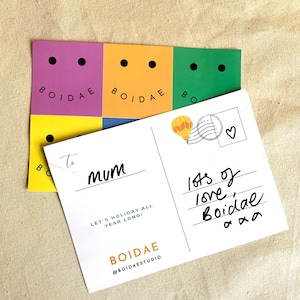 May include: A white postcard with a colorful hot air balloon illustration and the text "lots of love, Boidae" written in black ink. The postcard also has the text "To mum" and "Let's holiday all year long!" printed in black. The postcard is decorated with a sticker that says "BOIDAE" in black text on a yellow background.