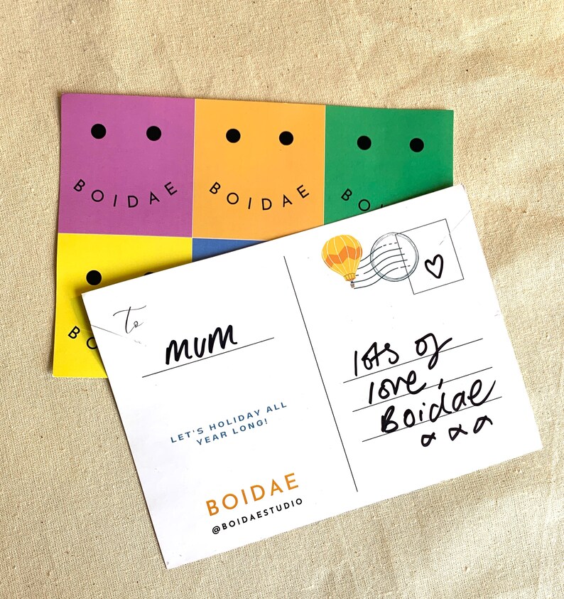 May include: A white postcard with a colorful hot air balloon illustration and the text "lots of love, Boidae" written in black ink. The postcard also has the text "To mum" and "Let's holiday all year long!" printed in black. The postcard is decorated with a sticker that says "BOIDAE" in black text on a yellow background.