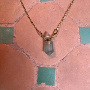 Pode incluir: Um colar de corrente de metal dourado com um pendente em forma de pequena garrafa de cristal transl&uacute;cido verde-p&aacute;lido, com uma tampa de cristal pontiaguda a condizer. A corrente liga-se &agrave; garrafa atrav&eacute;s de dois pequenos fechos lagosta.