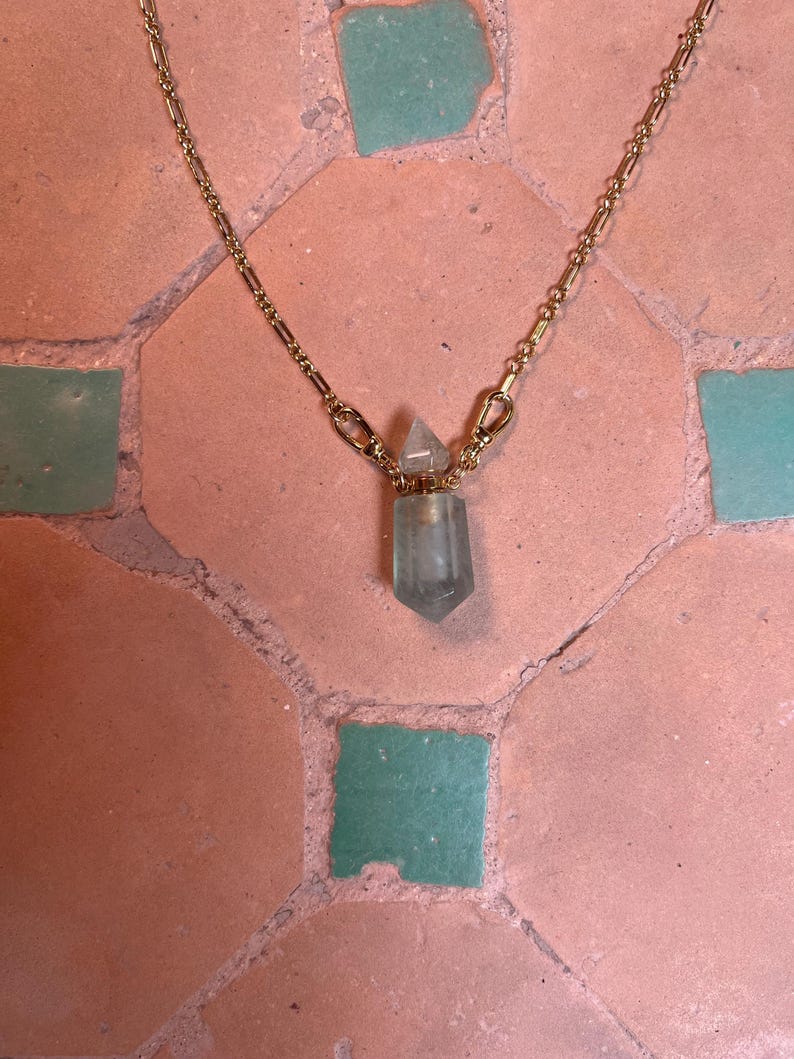 Pode incluir: Um colar de corrente de metal dourado com um pendente em forma de pequena garrafa de cristal transl&uacute;cido verde-p&aacute;lido, com uma tampa de cristal pontiaguda a condizer. A corrente liga-se &agrave; garrafa atrav&eacute;s de dois pequenos fechos lagosta.