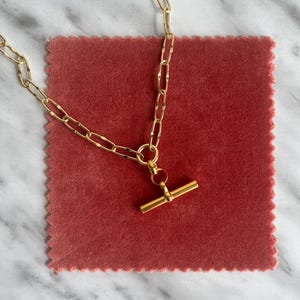 May include: A gold-colored necklace with a toggle clasp, resting on a square of red fabric. The chain links are elongated and textured. The background is a white and gray marble surface.
