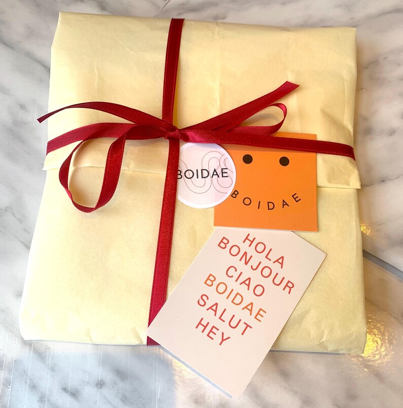 May include: A yellow gift box wrapped with a red ribbon and a white sticker with the word "BOIDAE" on it. The box is sitting on a white surface. A white card with the words "Hola Bonjour Ciao Boidae Salut Hey" is in front of the box.