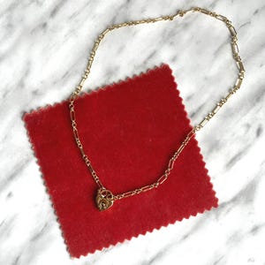 May include: A gold-colored necklace with a heart-shaped pendant featuring a keyhole design. The necklace is displayed on a red velvet surface, set against a white and gray marble background. The chain has a delicate, rectangular link design.