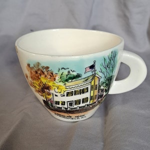 May include: White ceramic teacup with a hand-painted image of Abraham Lincoln's home in Springfield, Illinois. The image includes a yellow house with white windows, green trees, and an American flag.