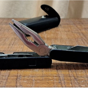 May include: A black multi-tool with pliers extended, showcasing various tools. The open tool reveals a silver blade and other implements. A black leather case sits in the background. The tool is on a wooden surface.