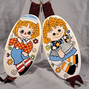 May include: Two oval-shaped, white ceramic wall hangings with a red-haired girl on each. The girl on the left is wearing a red and white checkered shirt, blue jeans, and red and white striped socks. The girl on the right is wearing a black and white dress with a white lace trim and orange and white striped socks. Both girls have black eyes and a red nose. The background is white with yellow flowers.