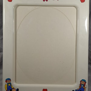 May include: White plastic picture frame with a red heart and a pink ribbon at the top. The frame has a white background with two cartoon dolls on the bottom, one boy and one girl.