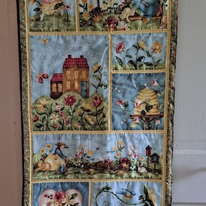 May include: A blue and yellow patchwork quilt with a floral theme. The quilt features several different scenes of gardens, flowers, and birds. The text on the quilt reads "Nurture my garden, Nurture my soul", "Where flowers bloom, so does hope", "The earth laughs with flowers", "The hum of bees is the song of the garden", "Gardeners use flowers as paint, soil and sky as canvas", "Love awaits at the edge of a petal", and "A garden should not be without bird songs".