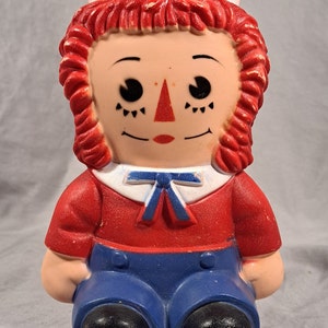 May include: A vintage plastic Raggedy Ann doll toy with red hair, a white sailor hat, a red shirt, blue pants, and black shoes.