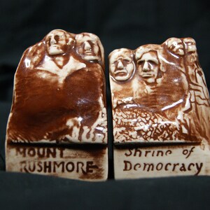 May include: A pair of brown and white ceramic bookends featuring the Mount Rushmore National Memorial. The left bookend reads "Mount Rushmore" and the right bookend reads "Shrine of Democracy".