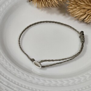 May include: A minimalist bracelet featuring a gray cord and a small, oval silver charm. The bracelet is displayed on a white, textured plate, with dried floral accents in the background. This simple design is suitable for everyday wear.