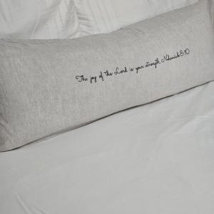 May include: A long, rectangular light gray pillow with the text "The joy of the Lord is your strength Nehemiah 8:10" in black script. The pillow is on a white bed.
