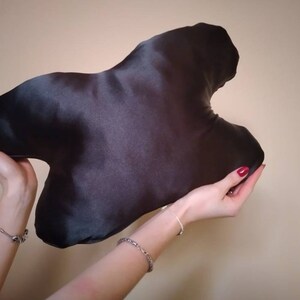 以下が含まれることがあります： 不規則な大きな抽象的な形をした黒いサテン製の旅行用枕。枕は人の手で持たれている。