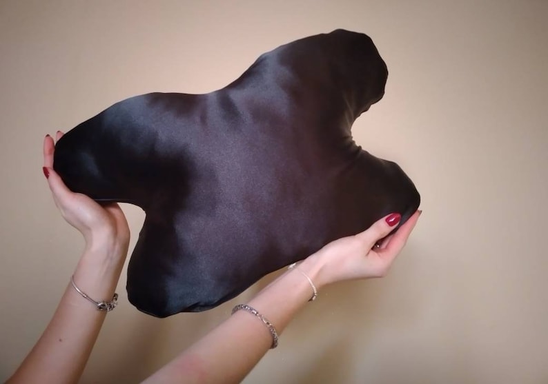 以下が含まれることがあります： 不規則な大きな抽象的な形をした黒いサテン製の旅行用枕。枕は人の手で持たれている。