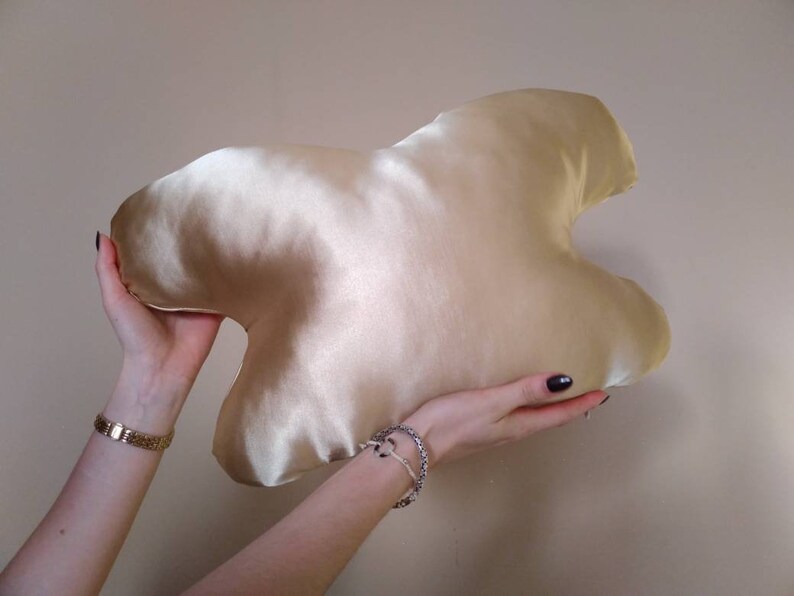 以下が含まれることがあります： スタイリッシュなハート型のゴールドサテンの枕。枕は両手で持たれています。