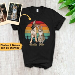 May include: Black t-shirt featuring a graphic of a dog and cat with a sunset background. The names "Buddy Felix" are printed below the animals. Two photos of the pets are shown above the shirt. Text on the left says "Photos & Names can be changed."