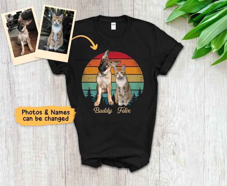 May include: Black t-shirt featuring a graphic of a dog and cat with a sunset background. The names "Buddy Felix" are printed below the animals. Two photos of the pets are shown above the shirt. Text on the left says "Photos & Names can be changed."