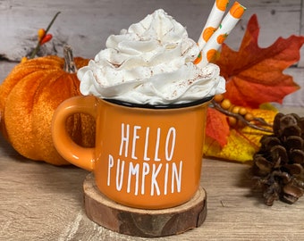 May include: An orange mug with the text "HELLO PUMPKIN" in white, filled with whipped cream and cinnamon. Two straws with pumpkin designs are in the mug. The mug sits on a wooden coaster, with a pumpkin and fall foliage in the background.