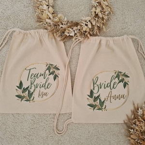 Könnte beinhalten: Zwei weiße Canvas-Rucksäcke mit goldenen und grünen Blumenmustern. Ein Rucksack sagt "Team Bride Lisa" und der andere sagt "Bride Anna".