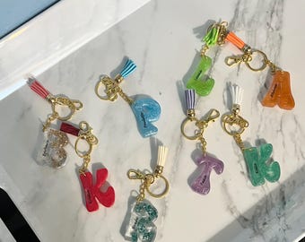 May include: A collection of 10 colourful resin letter keychains with gold key rings and tassels. The letters are K, R, T, L, A, B, F, J, and K. Each letter has a different colour and glitter embedded in the resin. The letters are spelled out as "Kristen, Barbara,  Renee,  Katie,  Lillie,  Andrew,  Fausto,  Jenna,  and  Killian."