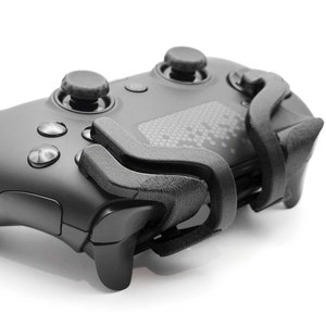 May include: A matte black video game controller featuring the word ENVISION printed on the side. The device includes two analog sticks, four action buttons, and a textured black plastic attachment secured over the right-hand shoulder triggers.