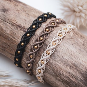 Könnte beinhalten: Drei geflochtene Armbänder mit goldenen Perlen auf einer Holzoberfläche. Die Armbänder sind schwarz, braun und cremefarben.