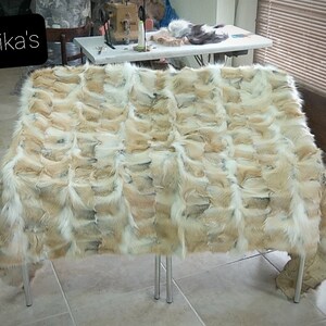 May include: A large, rectangular fur throw made from fox pelts. The fur is a mix of white, brown, and black, and the throw is laid out on a table with metal legs. The throw is made up of many individual pelts sewn together.