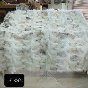 May include: A white fur throw blanket made of fox fur. The blanket is laid out on a table with a sign that says "Kika's" in black text.