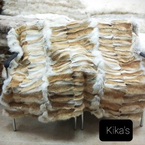 May include: A tan and white rabbit fur throw blanket is draped over a chair. The blanket is made of strips of fur sewn together. The blanket is labelled "Kika's".