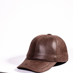 Puede incluir: Una gorra de béisbol de cuero marrón con una visera curvada y detalles de costura. La gorra está sobre una superficie blanca junto a un teléfono inteligente negro.