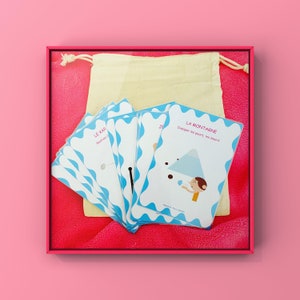 May include: A collection of illustrated cards with blue wavy borders and French text, and a beige drawstring bag. The cards display colourful illustrations and phrases such as "LA MONTAGNE" and "LE KAR". The set is presented within a pink frame.