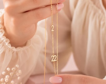 May include: A gold-toned necklace featuring a delicate chain with two suspended numeric charms. The upper charm is the number 2, and the lower charm is the number 22. The metal has a polished finish, and the design is simple and linear.