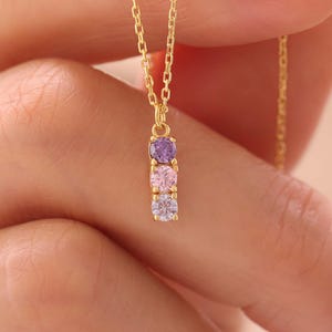 May include: A gold-colored necklace with three colorful gemstones. The gemstones are purple, pink, and white. The necklace is held up by a person's hand.