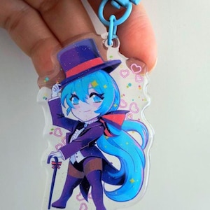 May include: An acrylic keyring featuring a cartoon character with long blue hair wearing a purple top hat, a purple tailcoat, and a white vest. The character holds a small cane and is surrounded by pink heart outlines and colourful confetti accents.