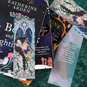 Puede incluir: Un marcapáginas con una mujer de pelo largo y oscuro, vestida con un vestido verde y blanco, sosteniendo un pájaro pequeño. El marcapáginas tiene un fondo azul y blanco con un diseño de copo de nieve y flores blancas. El texto "No hay monstruos en el mundo, ni santos. Solo infinitos matices tejidos en el mismo tapiz, luz y oscuridad. El monstruo de un hombre es el amado de otro. Los sabios lo saben." está impreso en el marcapáginas.