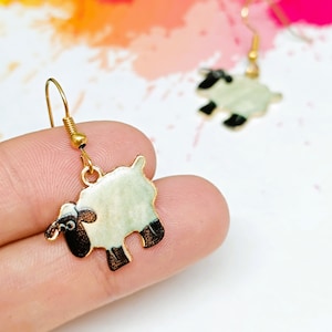 May include: A pair of gold tone earrings featuring a white sheep with black accents. The sheep are dangling from gold tone hooks.