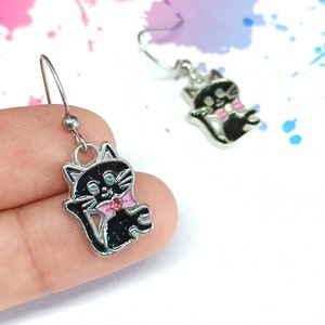May include: A pair of silver dangle earrings with black cat charms. The cats have pink bows and green eyes.