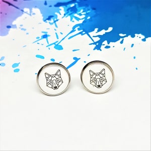 Könnte beinhalten: Ein Paar silberfarbene Ohrstecker. Jeder Ohrring hat ein weißes, kreisförmiges Gesicht mit einem schwarzen, geometrischen Wolfsdesign. Die Ohrringe sind vor einem weißen Hintergrund mit blauen Farbspritzern platziert.