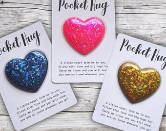 May include: Three pocket hug cards with a heart-shaped glitter filled resin charm. Each card has a different color charm: blue, pink, and gold. The cards have the text "Pocket Hug" and a message about love and hugs.