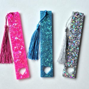 Op de afbeelding: Drie kleurrijke bladwijzers met hartvormige uitsparingen en kwastjes. De bladwijzers zijn gemaakt van hars en hebben een glinsterende afwerking. De kleuren zijn roze, blauw en zilver.
