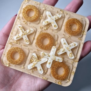 Op de afbeelding: Een wit en gouden tic-tac-toe spel met gouden glitter ingebed in de hars. De spelstukken zijn cirkels en X'en.