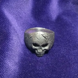May include: A silver skull ring with a textured surface. The ring is designed with a detailed skull face.