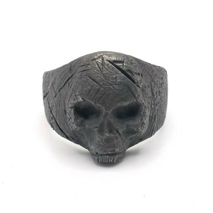 May include: A black skull ring with a textured surface. The skull is detailed with eye sockets, a nose, and a mouth.