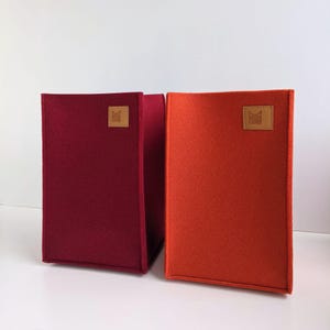 May include: Two rectangular felt storage bins, one in a deep burgundy and the other in a vibrant orange. Each bin has a square leather patch with a logo. The bins are designed to stand upright.
