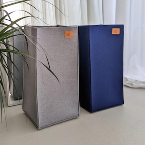 Può includere: Due contenitori rettangolari in feltro, uno grigio e uno blu navy. Ogni contenitore ha una toppa quadrata in pelle marrone con un logo. I contenitori sono su una superficie bianca vicino a una finestra con una pianta. Altezza dei contenitori: circa 60 cm.
